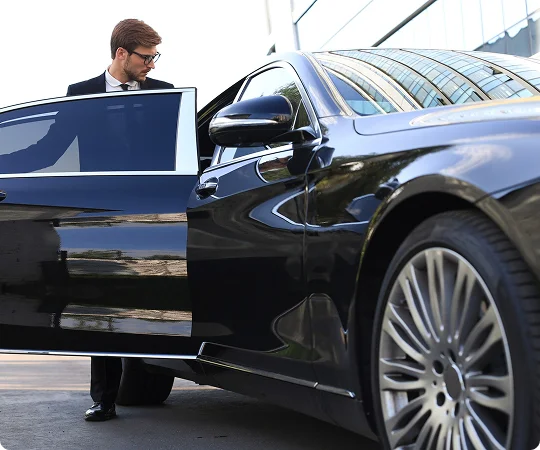 A well-dressed man steps out of a polished black luxury car, captured from a sleek low angle with sharp reflections on the vehicle's body.