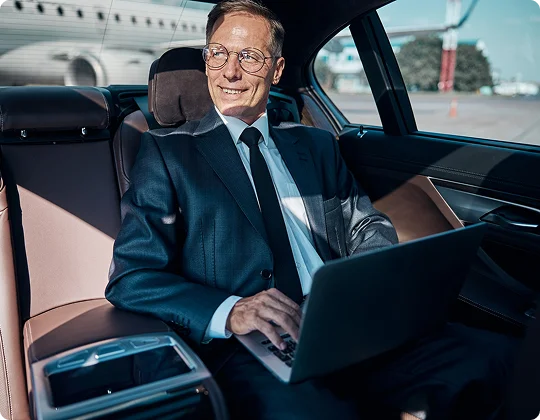 A business traveler works on his laptop in the backseat of a luxury car, with an airport runway scene visible through the window.