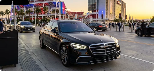 A black Mercedes-Benz sedan pulls up to a valet station in front of a large, brightly lit sports stadium at sunset.