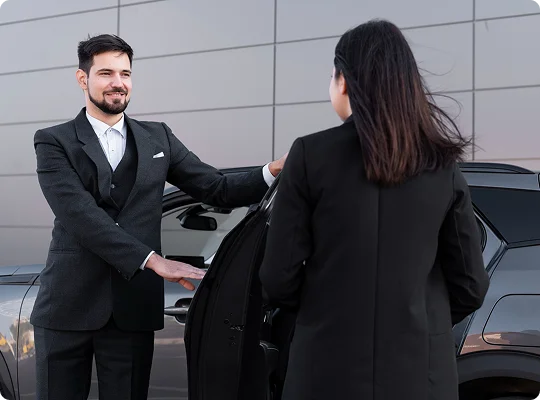 A professional chauffeur holds the car door open for a woman in formal attire, set against a sleek urban wall for a polished, service-focused impression.