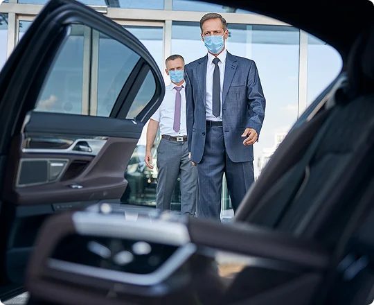 Business travelers in suits and face masks approach an open luxury car door, viewed from a spacious backseat angle, ready for a safe and professional pickup.