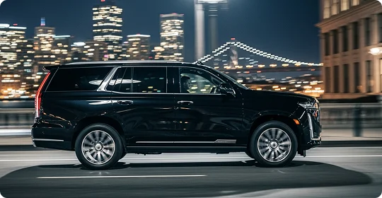 A black luxury SUV (appears to be a Cadillac Escalade) is driving quickly on a city street at night. The background shows a blurred cityscape with illuminated buildings and a large suspension bridge (likely the Bay Bridge in San Francisco) strung with lights.