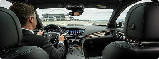Interior view from the back seat of a luxury car, showing a man in a suit driving on a highway toward a large stadium.