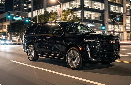 A black Cadillac Escalade SUV is driven quickly down a wide city street at night, with a motion blur effect. The background features brightly lit office buildings and streetlights.