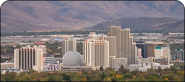 Aerial view of the downtown skyline of Reno, Nevada, featuring high-rise casinos and hotels, including the large silver geodesic dome structure of the Peppermill Resort, set against a backdrop of the dry, rolling mountains of the Great Basin.