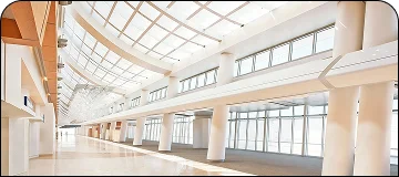 A wide-angle interior view of a modern, bright, and expansive airport or convention center terminal. The space features a high, curved glass roof and skylights that let in natural light, white support columns, and large windows along the wall. The floor is light-colored and polished, and the space appears mostly empty.