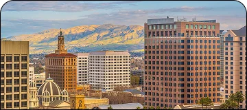 A daytime view of a modern downtown skyline, likely in the San Francisco Bay Area (potentially Oakland or San Jose). Prominent multi-story office buildings of various colors (brown, beige, white) fill the foreground, including one with a domed structure at its base and another with a distinctive older, brick tower in the center-left. Rolling, grassy golden hills are visible in the background under a partly cloudy blue sky.