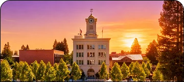 A horizontal, wide-angle photo of a historic-looking, light-colored, multi-story building with a central clock tower and weathervane. The scene is dramatic due to a vibrant sunset sky with gradients of orange and purple, framed by lush green trees in the foreground and sides.