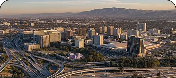 Aerial panoramic view of Silicon Valley California, showcasing the skyline of mid-rise buildings and complex highway interchanges in the foreground, all set against a backdrop of hazy mountains in the distance during sunset.
