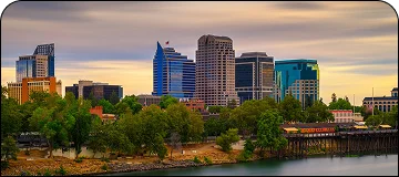 A horizontal photograph of the Sacramento, California, downtown skyline at sunset, featuring several modern high-rise buildings towering above a foreground of lush green trees and parks.