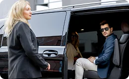A male passenger wearing sunglasses smiles as he steps out of a large black van or SUV. A female chauffeur or assistant is standing outside the vehicle's open door.