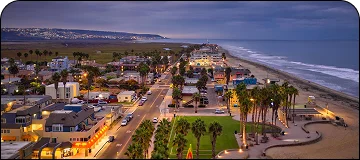 An elevated, twilight view of a coastal town, with a street lined by palm trees and illuminated buildings running parallel to a sandy beach and the ocean.