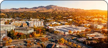 An aerial view of a city neighborhood during autumn, showing a mix of commercial buildings, residential areas, and streets surrounded by dense trees with bright fall foliage (red, orange, and yellow). The sunlight is intensely bright on the right side of the image, and a chain of large, dry mountain hills is visible across the background.