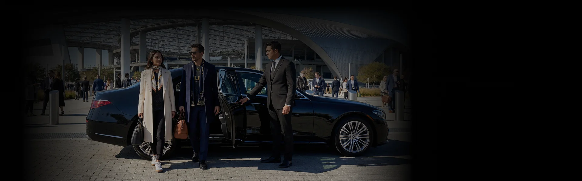 A well-dressed couple arriving at a stadium in a black luxury sedan, with a chauffeur holding the door open. Background shows outside view of a stadium.