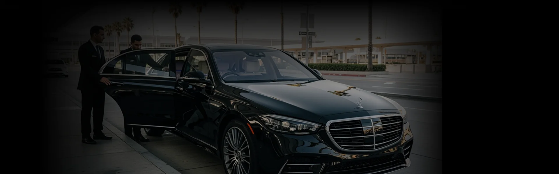 A chauffeur in a suit holds open the door of a black sedan for a passenger on an airport drop-lane.