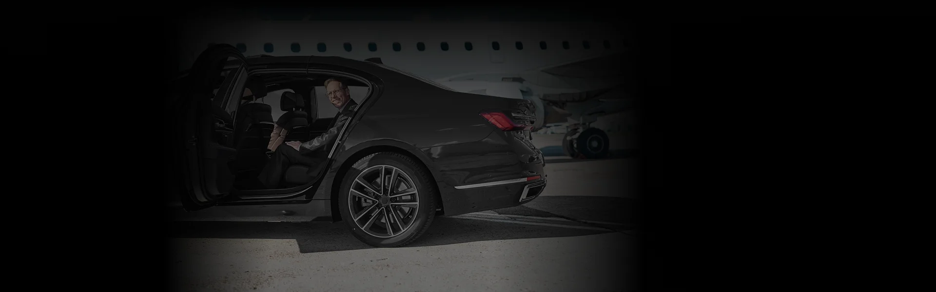 Chauffeur opens luxury black sedan at tarmac with passenger smiling inside, angled side-rear view for Oakland Airport Car Service.