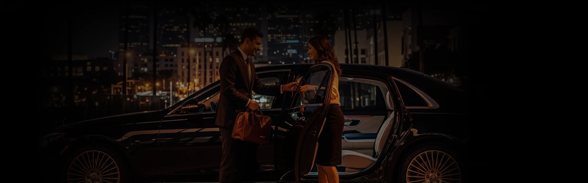 A chauffeur in a suit helps a female passenger with her luggage at night next to a black sedan.