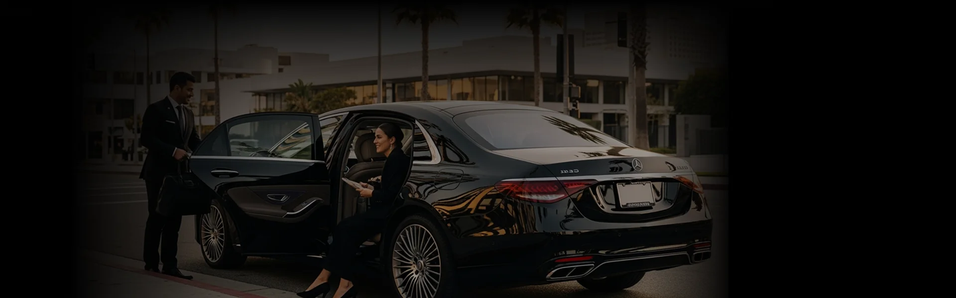 A chauffeur in a suit holds a bag while a female passenger gets out of a black sedan.