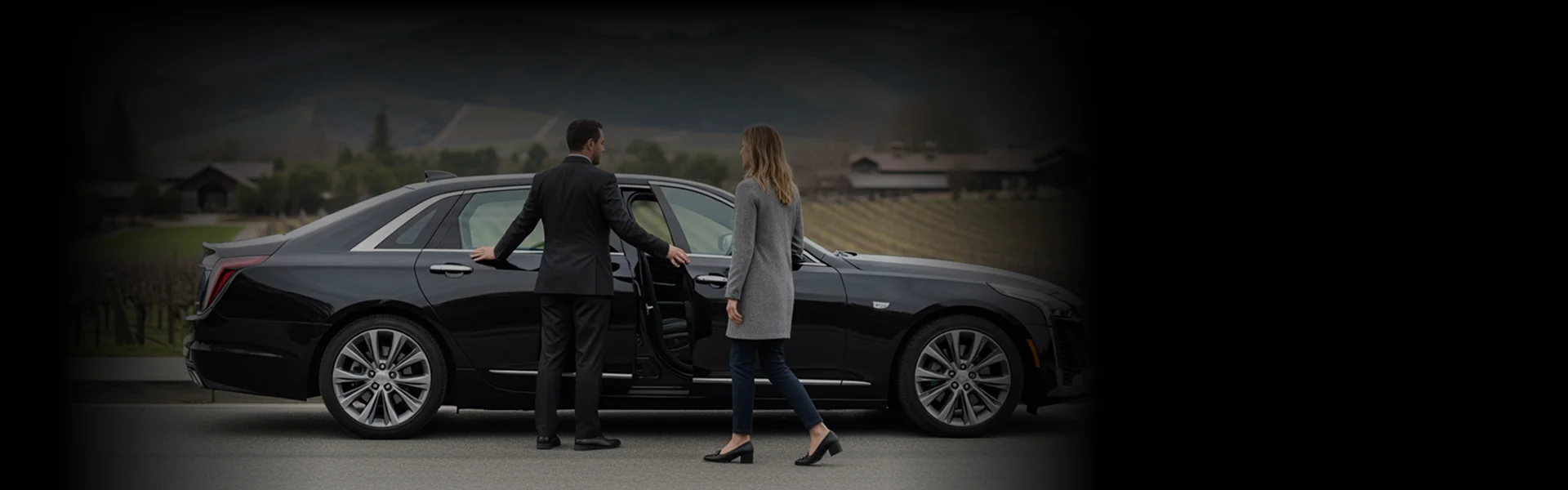 A chauffeur in a dark suit stands facing and holding the rear passenger door of a black luxury sedan open for a female passenger wearing a light grey coat. They are standing in an upscale, rural setting, with rolling hills and cultivated fields visible in the blurred background.