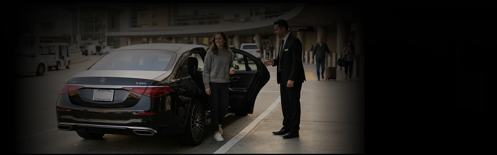 Chauffeur in a dark suit holding open the rear door of a black Mercedes-Benz S580 sedan for a woman in a grey sweater and dark pants who is stepping out of the car.