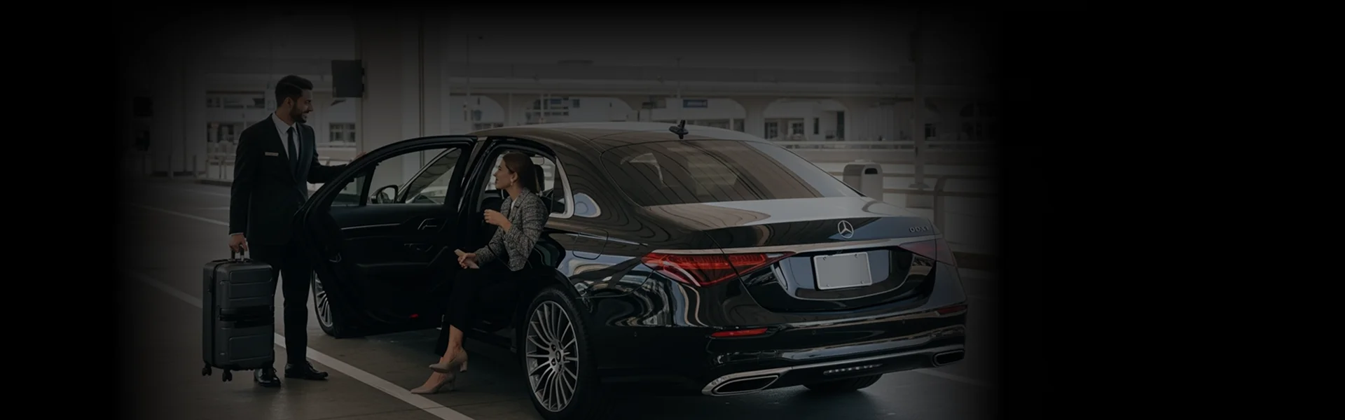 A chauffeur assists a female client exit a black business sedan while holding her luggage and rear door for her at the airport curbside.