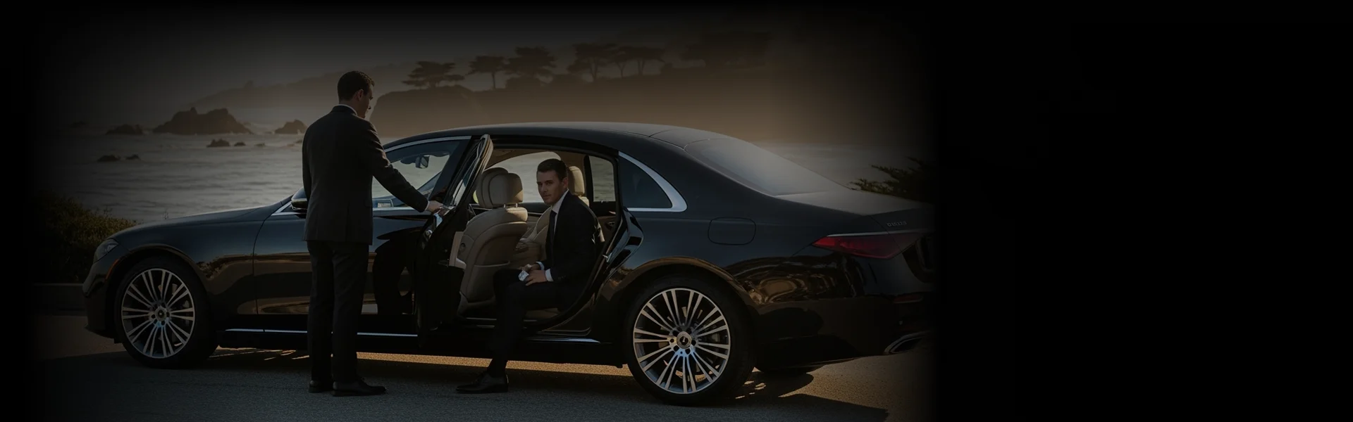 A chauffeur in a black suit opens the rear door of a luxury black sedan for a male passenger, also in a black suit, who is sitting inside. The scene is outdoors with a blurred background of a rocky coastline and the ocean at dusk or sunset. The image is dark and moody, highlighting the formal attire and the car's sleek design.