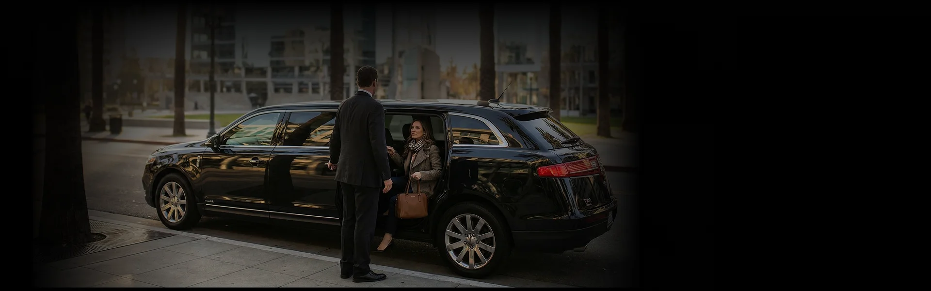 A decently dressed middle-aged woman steps out of a stretch limousine holding her brown handbag while a suited chauffeur hold the rear door opened for her on a street with palm trees and under-construction buildings loosely visible in the background.